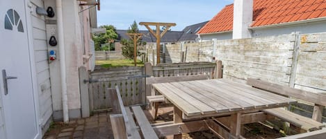 An outdoor patio area with a wooden table and benches, a white door with a security device, and a red-tiled roof visible in the background.