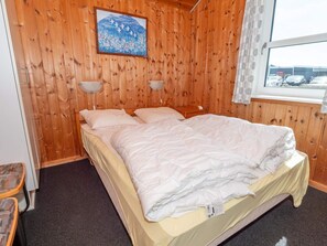 A room with wooden walls, a bed with white bedding, two bedside lamps, a window with a view of parked cars, and a painting on the wall. - 4 Star Holiday Home in Thisted (Thisted)