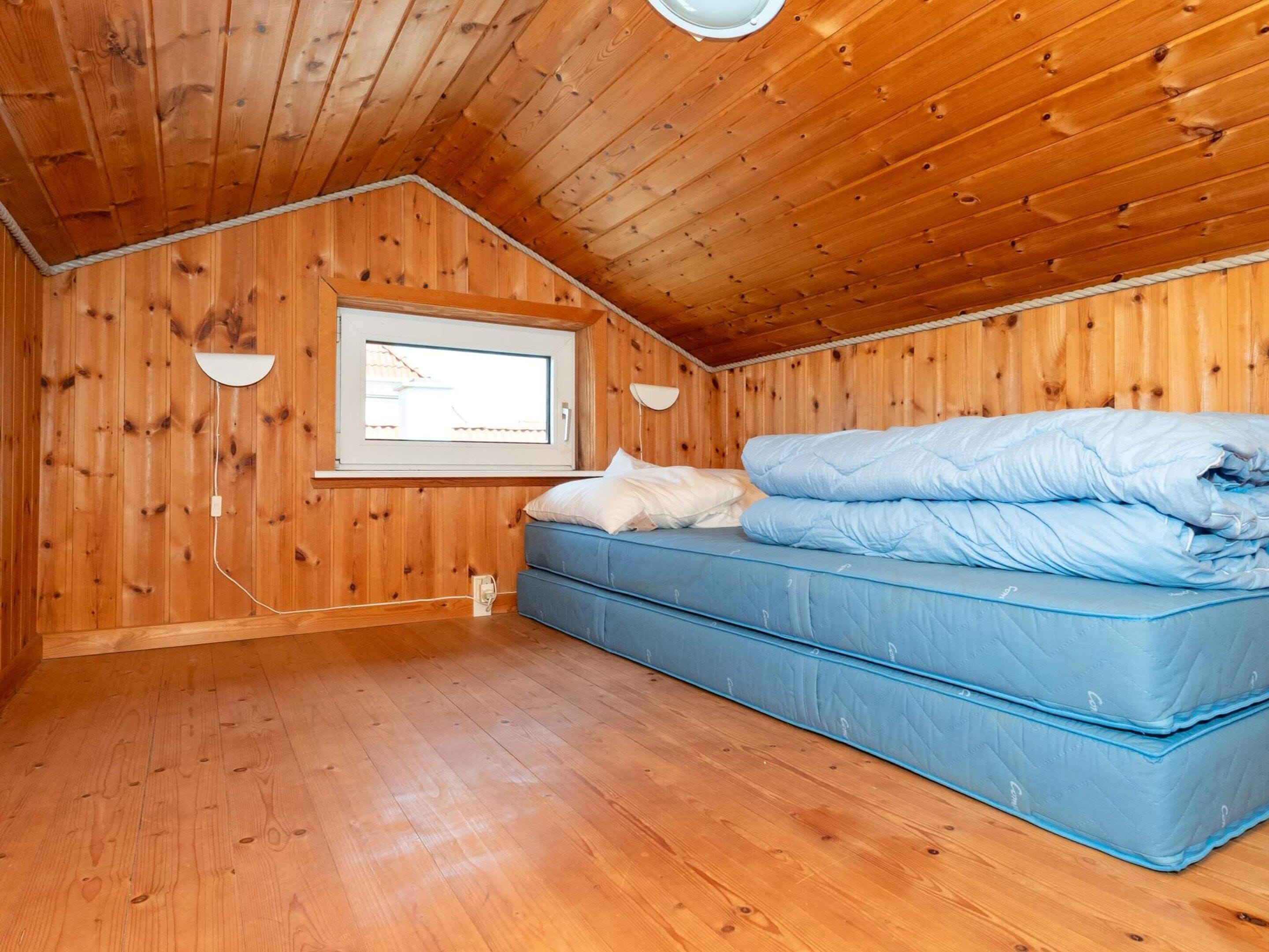 Una habitación pequeña con paneles de madera, una cama individual, una ventana y una lámpara fijada a la pared.