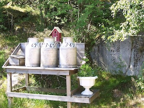 A wooden cart with three large white containers labeled with numbers, a small red birdhouse, and a white urn on a wooden platform in a wooded area. - 5 Person Holiday Home in Kungshamn (Kungshamn)