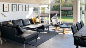 A modern living room with a black leather sofa, a wooden dining table, and a large window offering a view of the outdoors. - Wooden Holiday Home in Falster near Sea (Vaeggerlose)