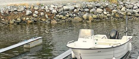 A boat docked at a wooden pier with a rocky shoreline and a house in the background.