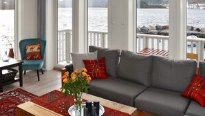 A living room with a sofa, a chair, a table with a vase of flowers, and large windows overlooking a body of water. - 5 Star Holiday Home in Brattvag (Ålesund)