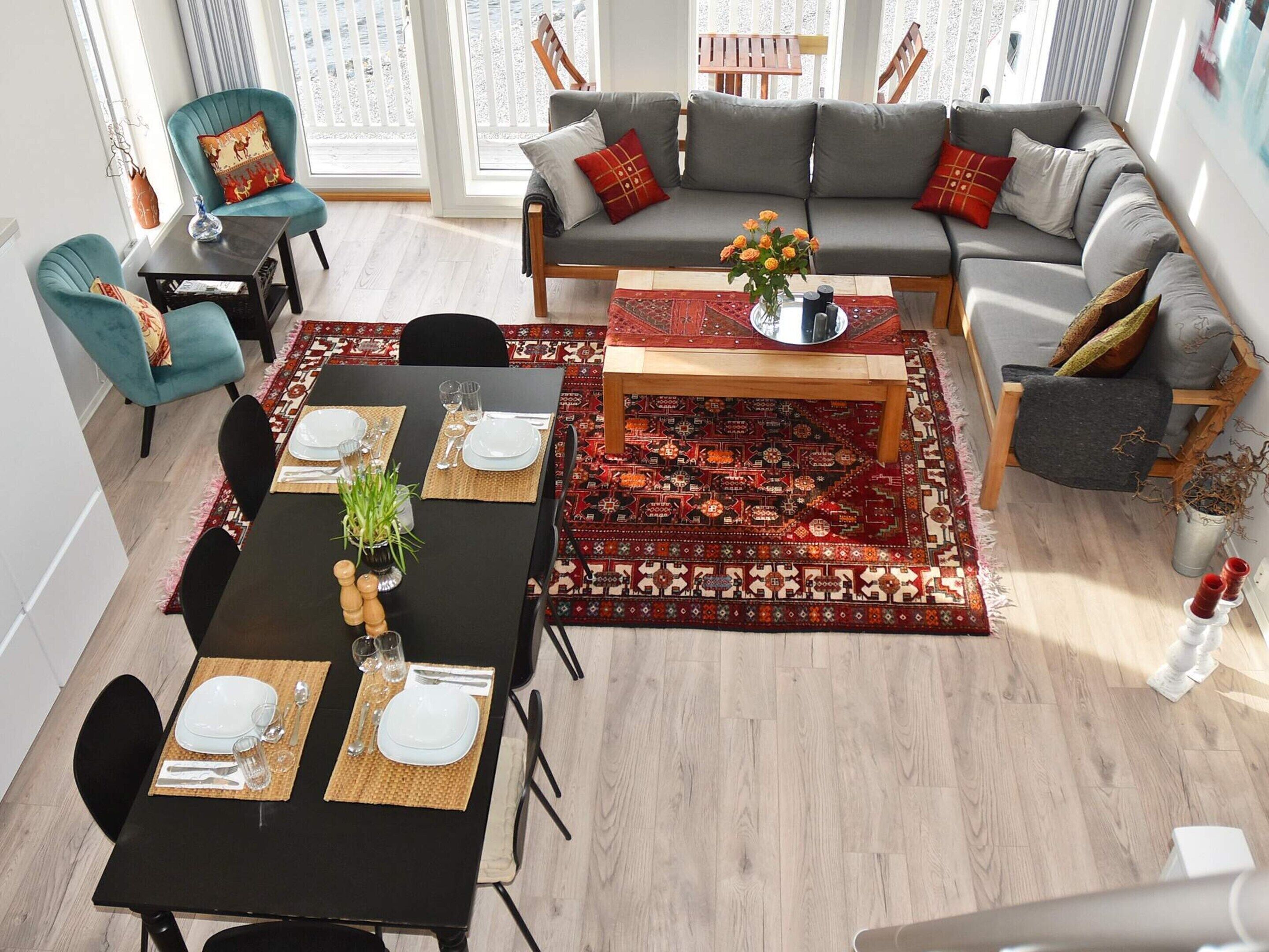 A modern living room with a dining area, featuring a grey sofa set, teal armchairs, a wooden coffee table, and a patterned rug.