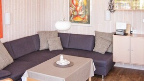 A room with a dark-colored sofa, a small table with a white tablecloth, a painting on the wall, and a cabinet with glassware. - Holiday Home in Otterndorf (Otterndorf)