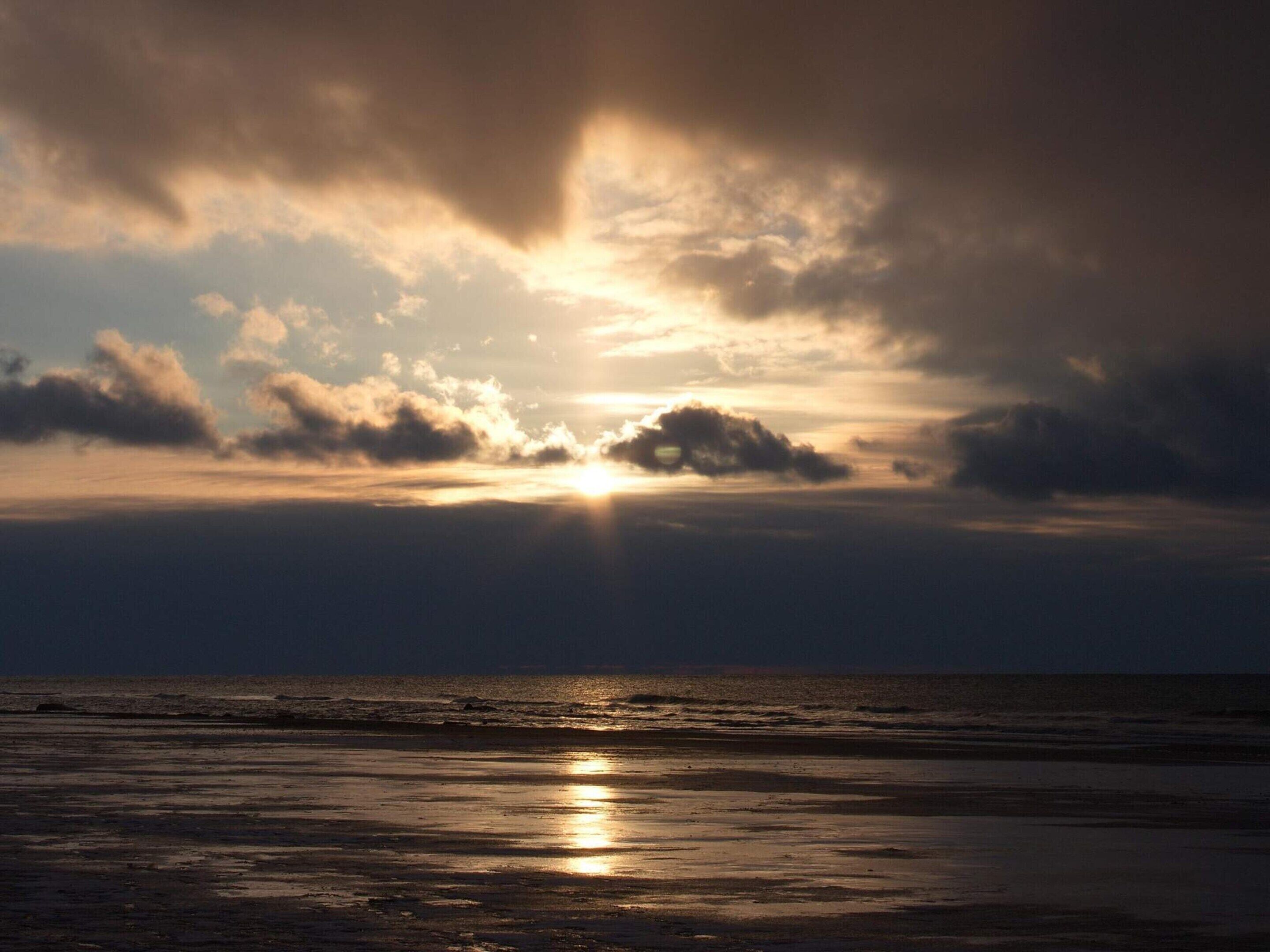 Uma praia com o sol se pondo atrás das nuvens, criando um efeito silhueta.