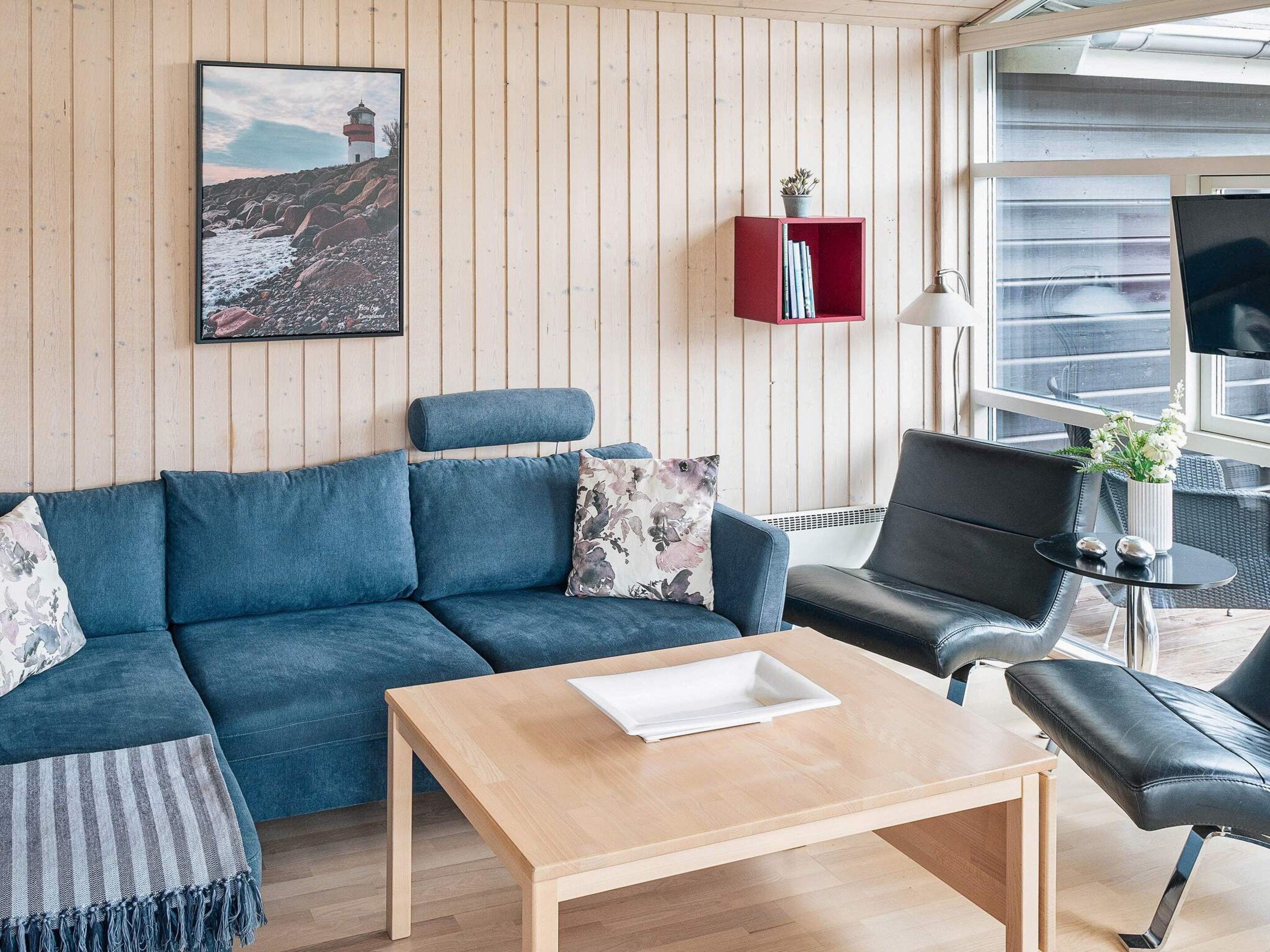 A modern living room with a blue sofa, a wooden coffee table, a black leather chair, and a red bookcase against a wooden wall.