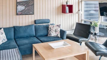 A modern living room with a blue sofa, a wooden coffee table, a black leather chair, and a red bookcase against a wooden wall.