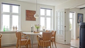 A dining area with a round table, six wooden chairs, a pendant light, and large windows. - Quaint Holiday Home in Bornholm near Sea (Svaneke)
