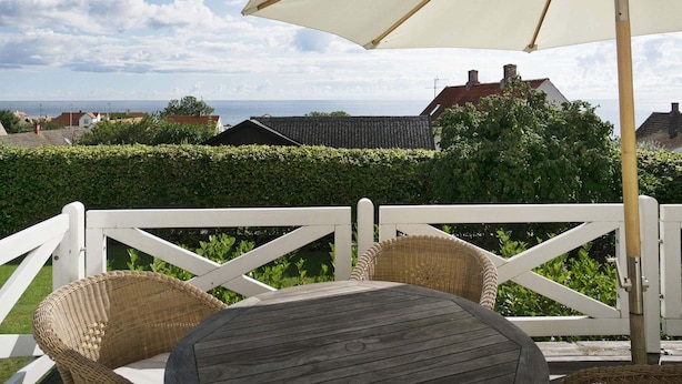 A wooden deck with a round table and wicker chairs, overlooking a sea view with houses and a hedge in the background. - Quaint Holiday Home in Bornholm near Sea (Svaneke)