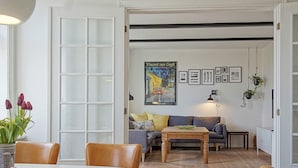 A modern living room with a large window, a sofa, a coffee table, and a dining table. - Quaint Holiday Home in Bornholm near Sea (Svaneke)