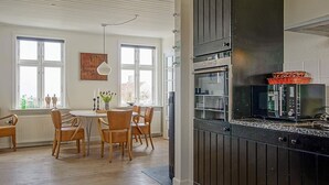 A modern kitchen with dark cabinetry, a microwave, and a built-in oven. There is a dining area with wooden chairs and a table, and large windows providing natural light. - Quaint Holiday Home in Bornholm near Sea (Svaneke)