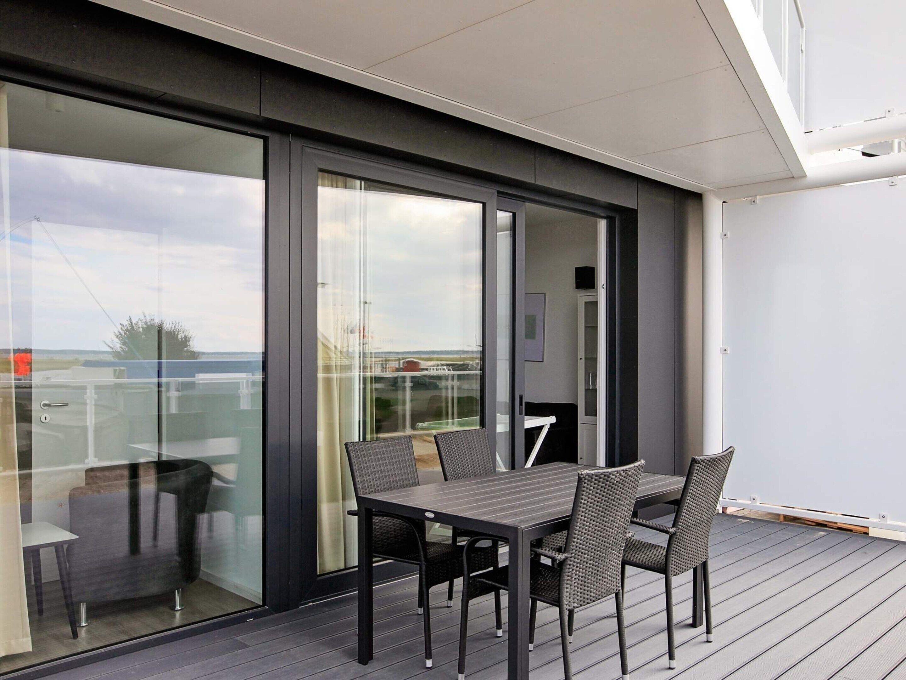 A modern outdoor dining area with a table and chairs, a glass door leading to a balcony, and a view of the sky and distant landscape.