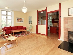 Un salon avec un canapé rouge, une table basse en bois et un fauteuil à bascule. On peut apercevoir une cuisine à travers une porte ouverte.