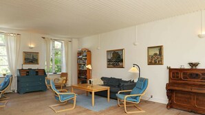 A living room with a wooden floor, a blue sofa, two blue armchairs, a wooden coffee table, a bookshelf, and a wooden cabinet. - Vintage Holiday Home in Syddanmark With Private Whirlpool (Sydlangeland)