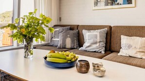 Una sala de estar moderna con un sofá, una mesa de centro con frutas y objetos decorativos, un jarrón con flores y un cuadro en la pared.