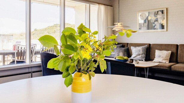 Una sala de estar moderna con una mesa redonda blanca, un jarrón amarillo y blanco con hojas verdes y flores amarillas, un sofá marrón con cojines, y un ventanal con vistas a la vegetación.
