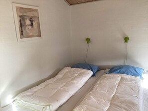 Ein schlichter Raum mit zwei Einzelbetten, jedes mit einem blauen Kopfkissen und einer weißen Daunendecke. An jeder Seite des Bettes befinden sich jeweils eine grüne Wandlampe.