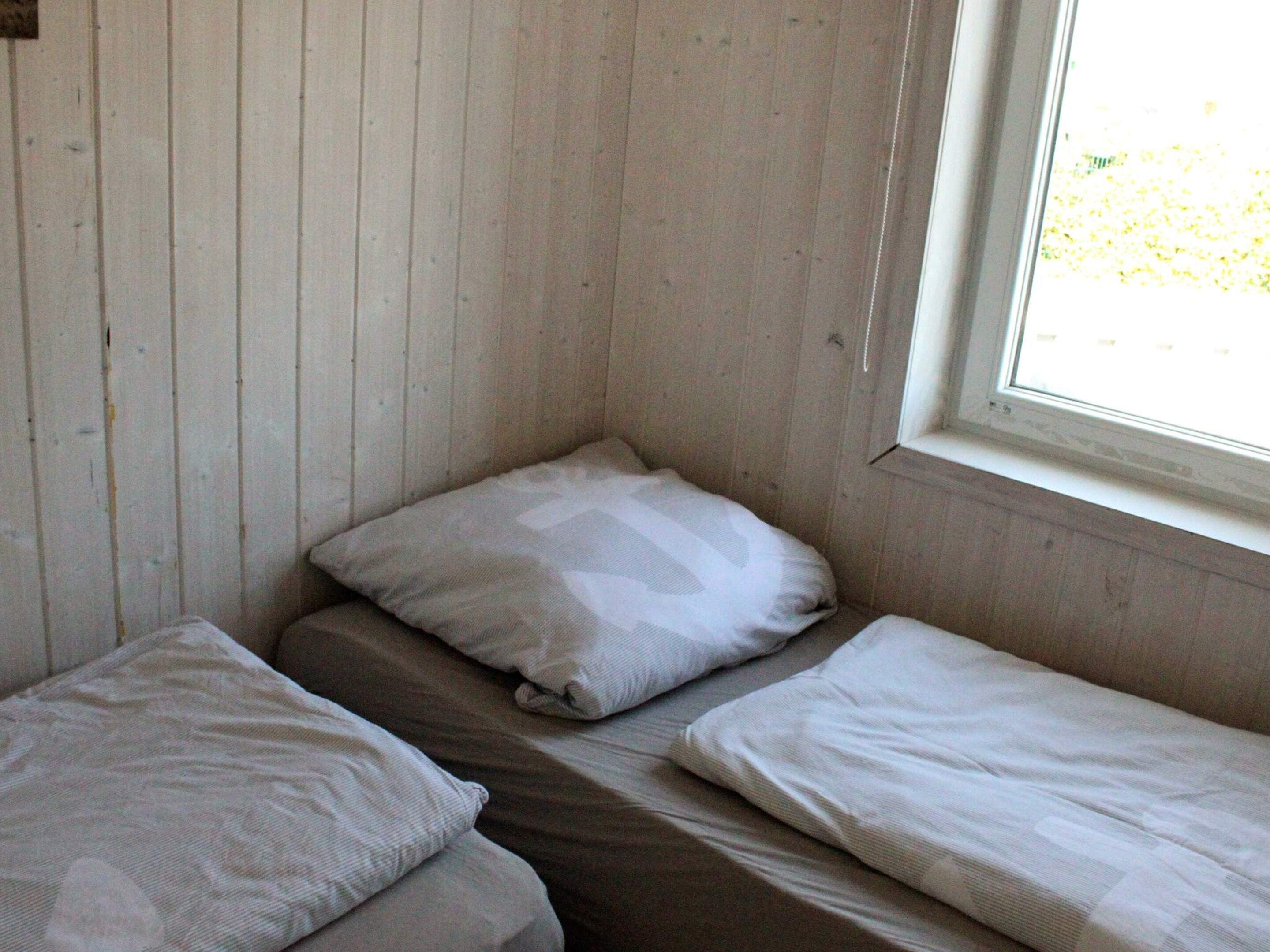 A small, single-bed room with a window, wooden walls, and a simple bed with two pillows.