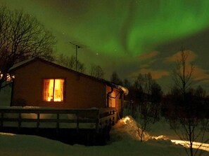 Une cabane avec une fenêtre bien visible, un sol recouvert de neige et un ciel nocturne dégagé.