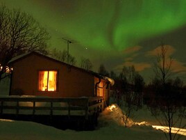 Una cabaña con una ventana visible, suelo cubierto de nieve y cielo nocturno despejado.