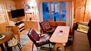 L’intérieur chaleureux d’un chalet, avec un décor en bois, une table à manger, des fauteuils en cuir, un canapé, un téléviseur fixé au mur et une fenêtre agrémentée de rideaux.