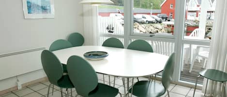 A dining area with a round table and six chairs, a window with a view of parked cars and houses, and a white radiator on the wall.