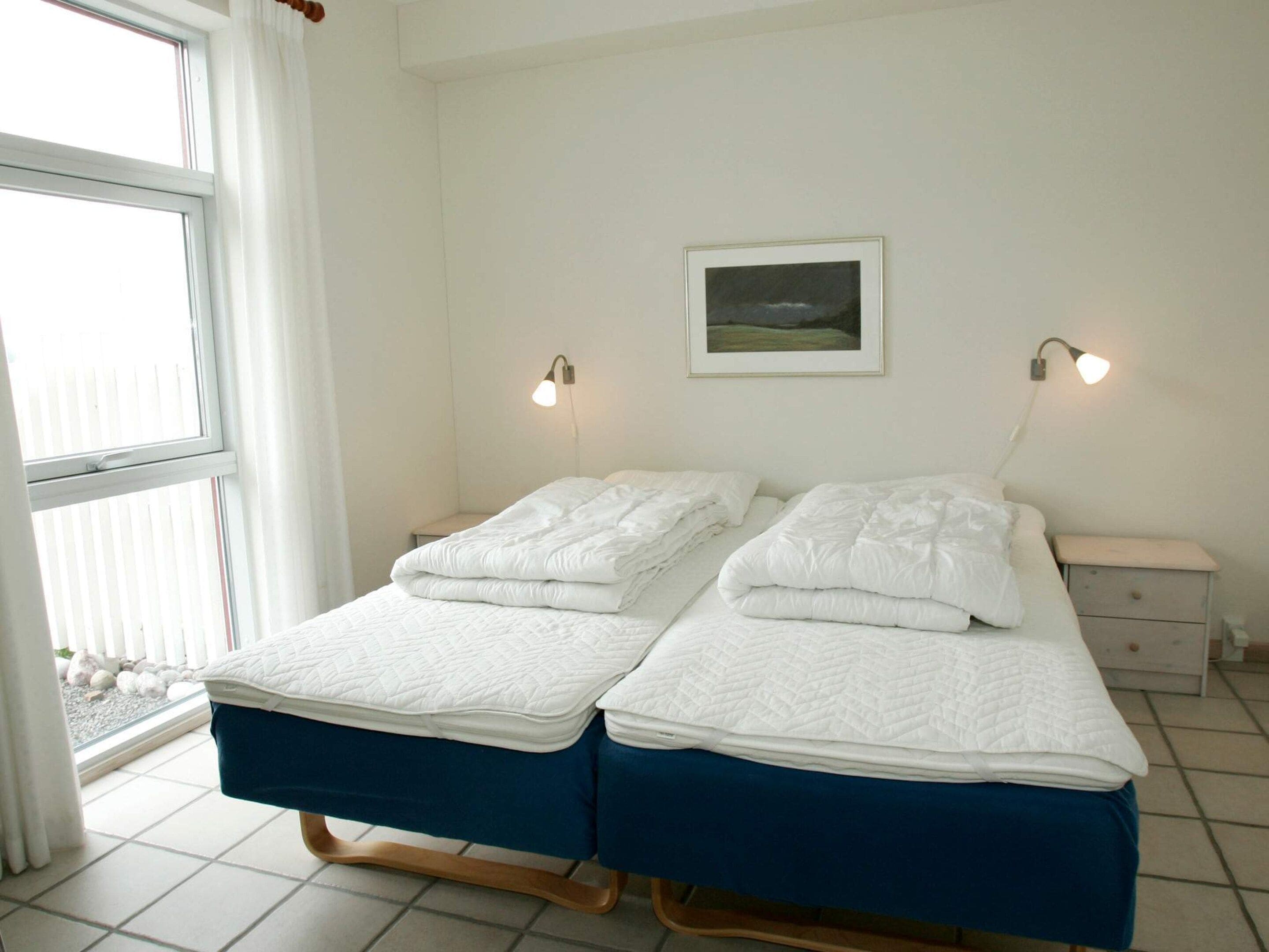 Cama doble con colchón azul y ropa de cama blanca, dos mesitas de noche, un cuadro colgado en la pared y una ventana con cortinas.