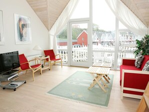 A room with a television, red chairs, a small table, a green rug, and a view of houses through large windows. - Quaint Holiday Home in Hasle near Sea (Hasle)