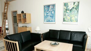 Ein Wohnzimmer mit einer schwarzen Ledersofa, einem Holztisch, einem gemusterten Teppich, einem Bücherregal und zwei abstrakten Bildern an der Wand.