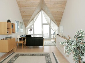 Quarto com teto inclinado, sofá preto, armário de madeira, mesa com abajur e uma janela grande com cortinas.