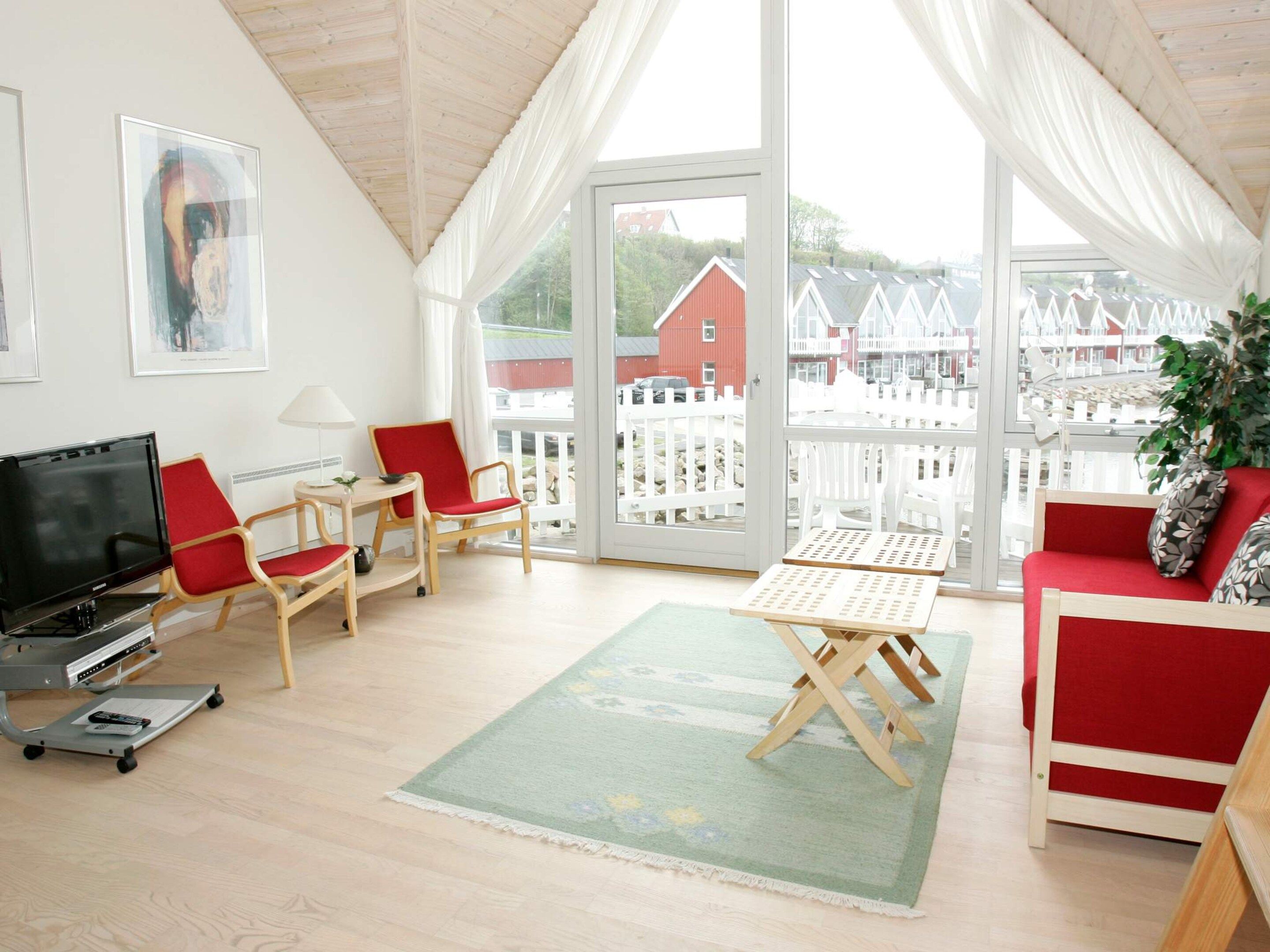 A room with a television, two red chairs, a small table, a sofa, and a view of houses through large windows.