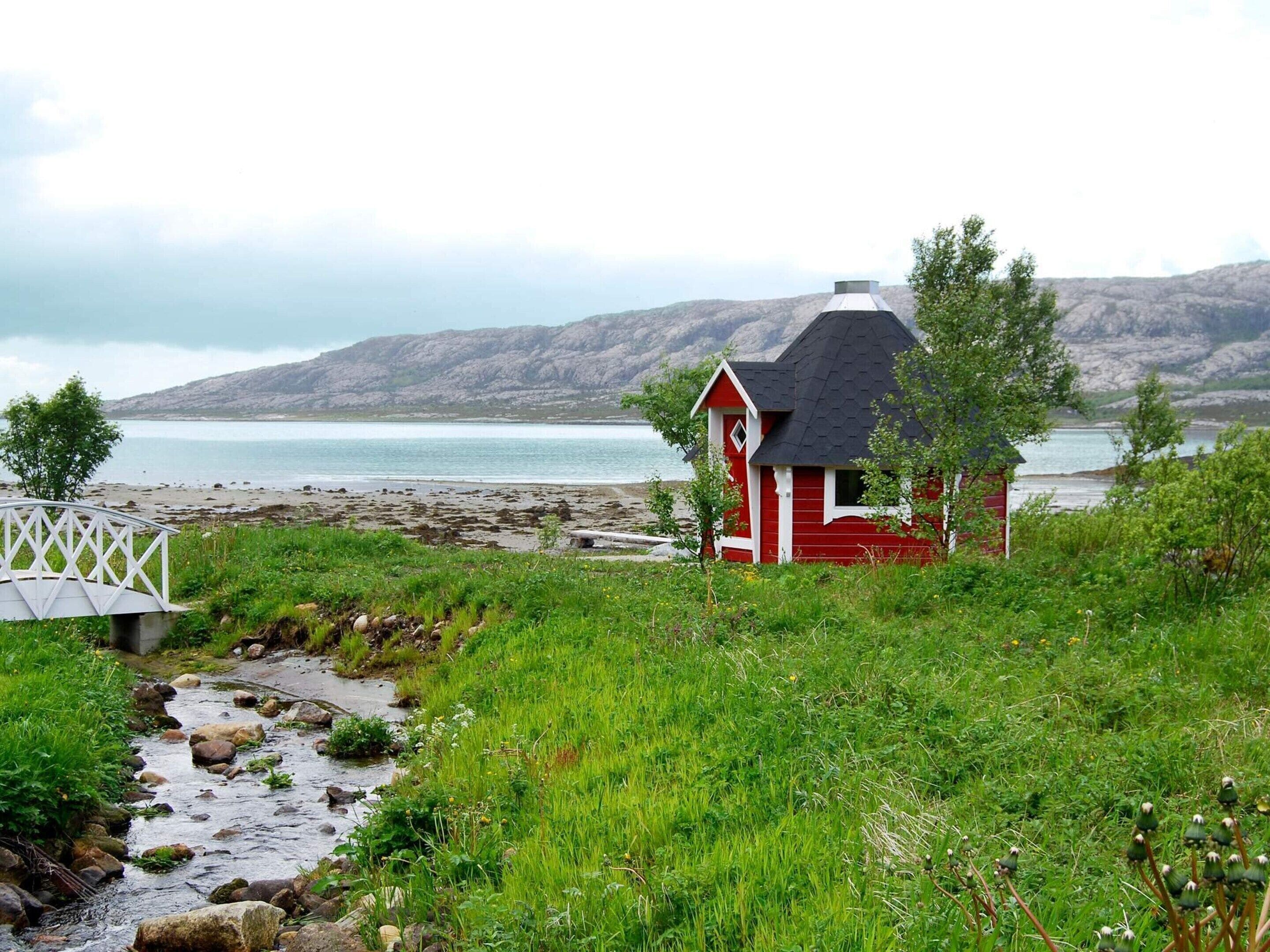 A red wooden structure with a white door and window, situated on a grassy area with a rocky shoreline and a bridge in the foreground.