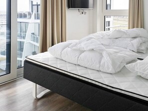 A modern hotel room with a large bed, a flat-screen TV mounted on the wall, and a view of the city through the window.