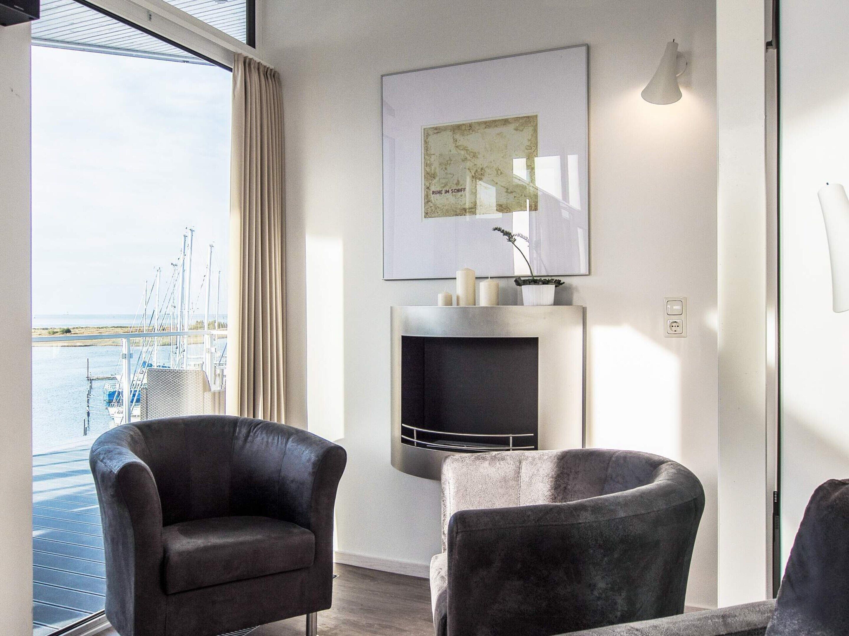 A modern living room with a large window overlooking a marina, two leather armchairs, a fireplace with decorative items, and a framed map on the wall.