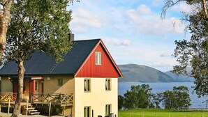 A two-story house with a red roof and beige walls, located near a lake with mountains in the background. - 6 Person Holiday Home in Alsvag-by Traum (Oksnes)