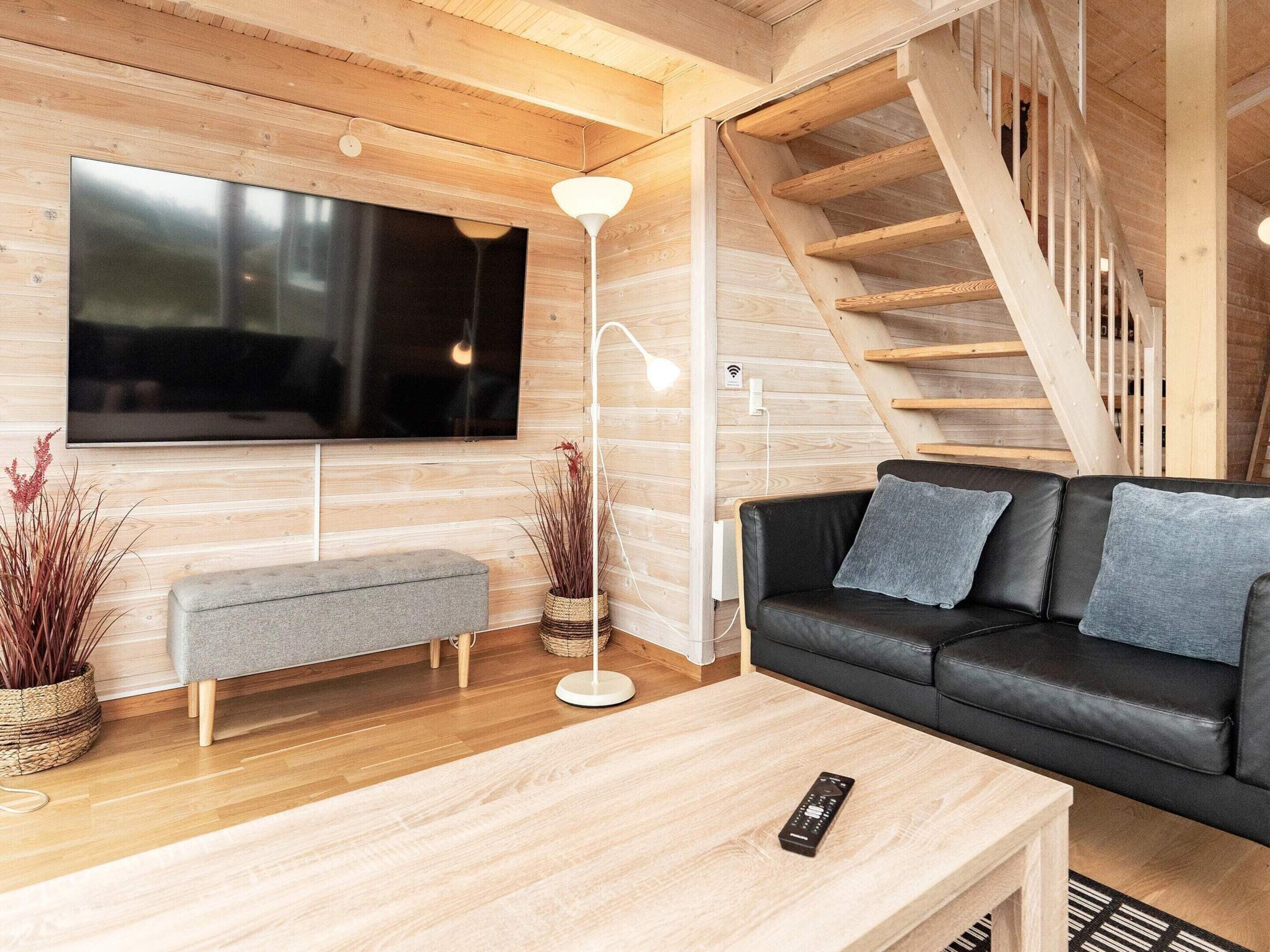 A modern living room with a flat-screen TV, a black leather sofa, a wooden coffee table, and a staircase.