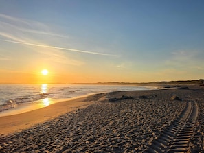 A beach with footprints leading to the water, a clear sky, and the sun setting over the horizon.