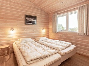 A wooden cabin bedroom with a bed, a small table, a wall-mounted light, and a window with curtains.