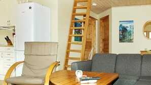 Un espacio habitable pequeño con kitchenette, una escalera de madera, un sofá gris y una mesita con un control remoto.