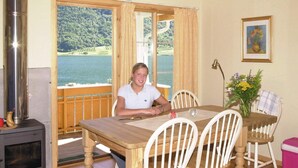 Una habitación con una mesa de madera, sillas, una estufa, un ventanal con vistas a un lago y un bosque, y un cuadro en la pared.