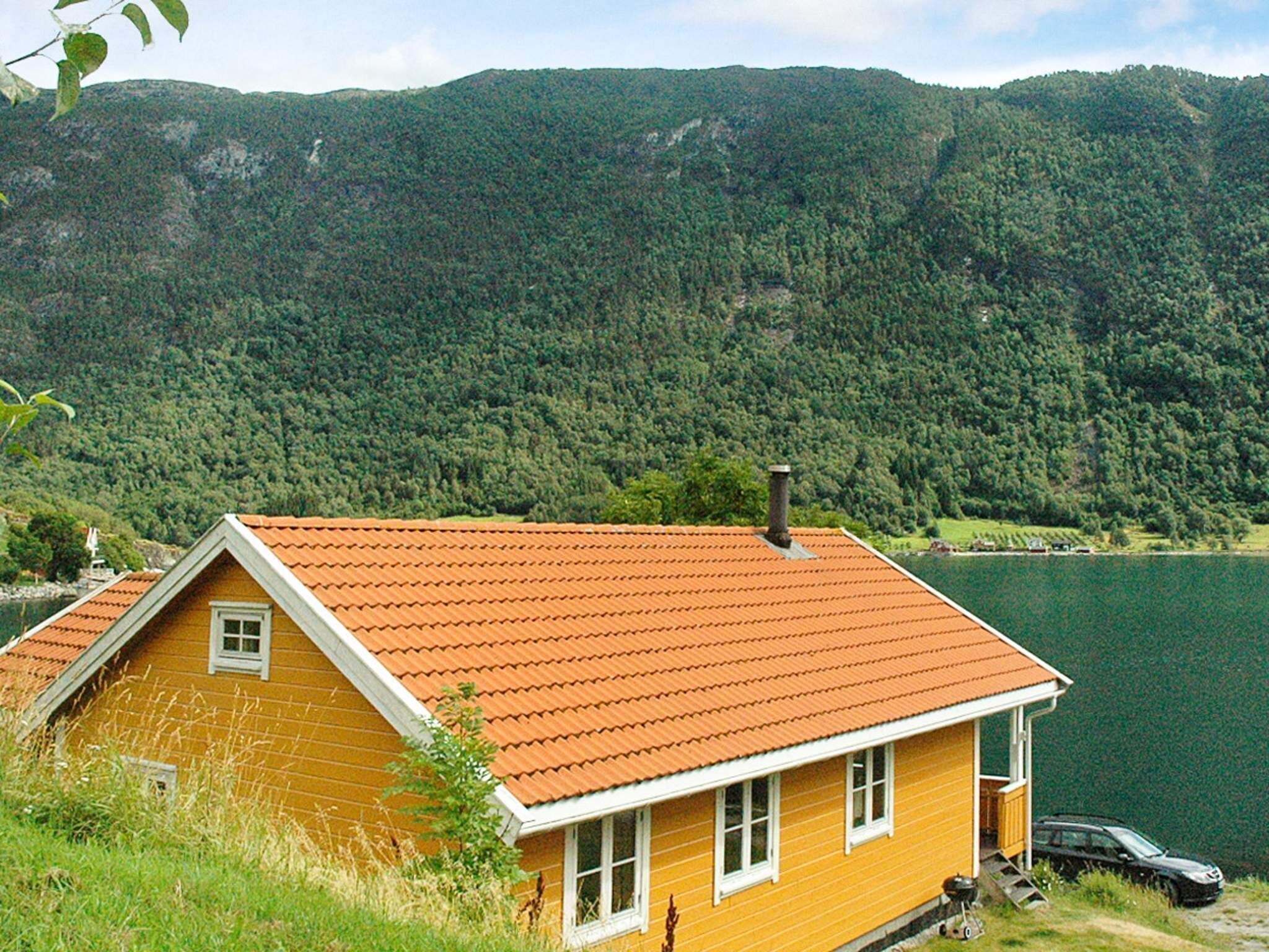 Une maison jaune avec un toit rouge, entourée d’un lac et de collines boisées.