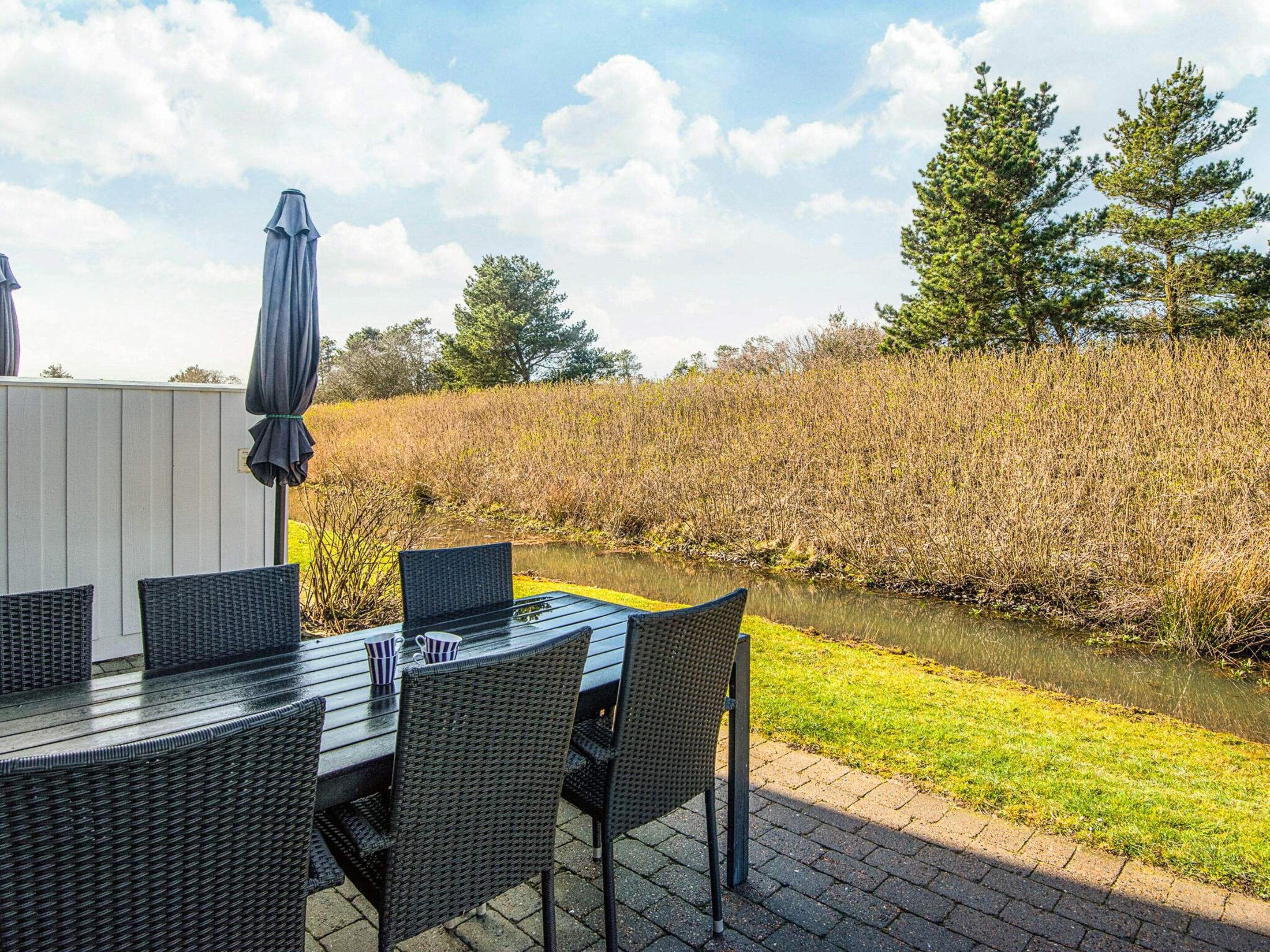 An outdoor dining area with a table and chairs, a covered umbrella, and a view of a water body and grassy field.