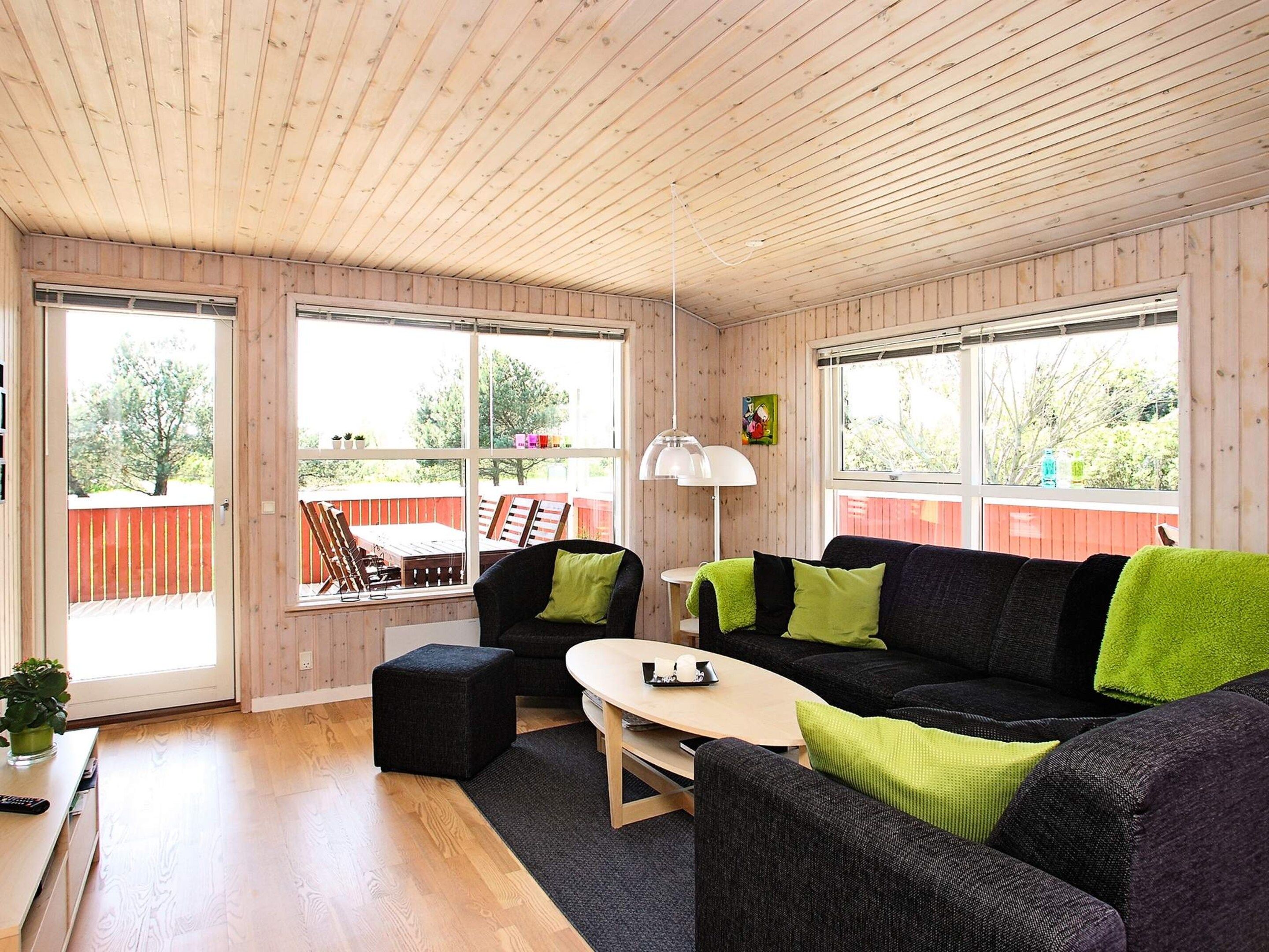 A cozy room with a wooden ceiling, a black sofa with green cushions, a small table, and a window overlooking a red deck.