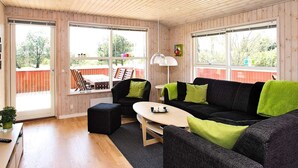 Ein gemütliches Wohnzimmer mit Holzbalkendecke, großen Fenstern, einem schwarzen Sofa mit grünen Kissen, einem kleinen Tisch und einem Sessel.