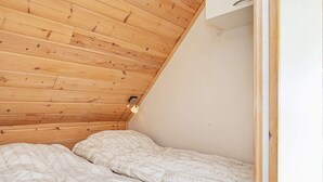 Una habitación pequeña y acogedora con techo inclinado y paneles de madera. Se observa una cama con una colcha estampada y una lámpara de noche.