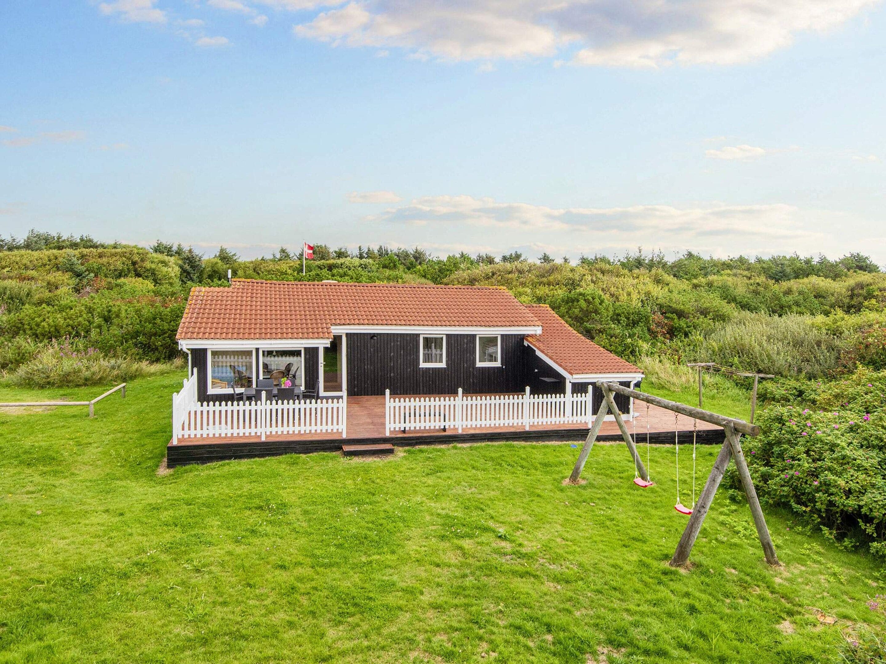 A two-story house with a red-tiled roof, a white picket fence, and a swing set in a grassy yard.