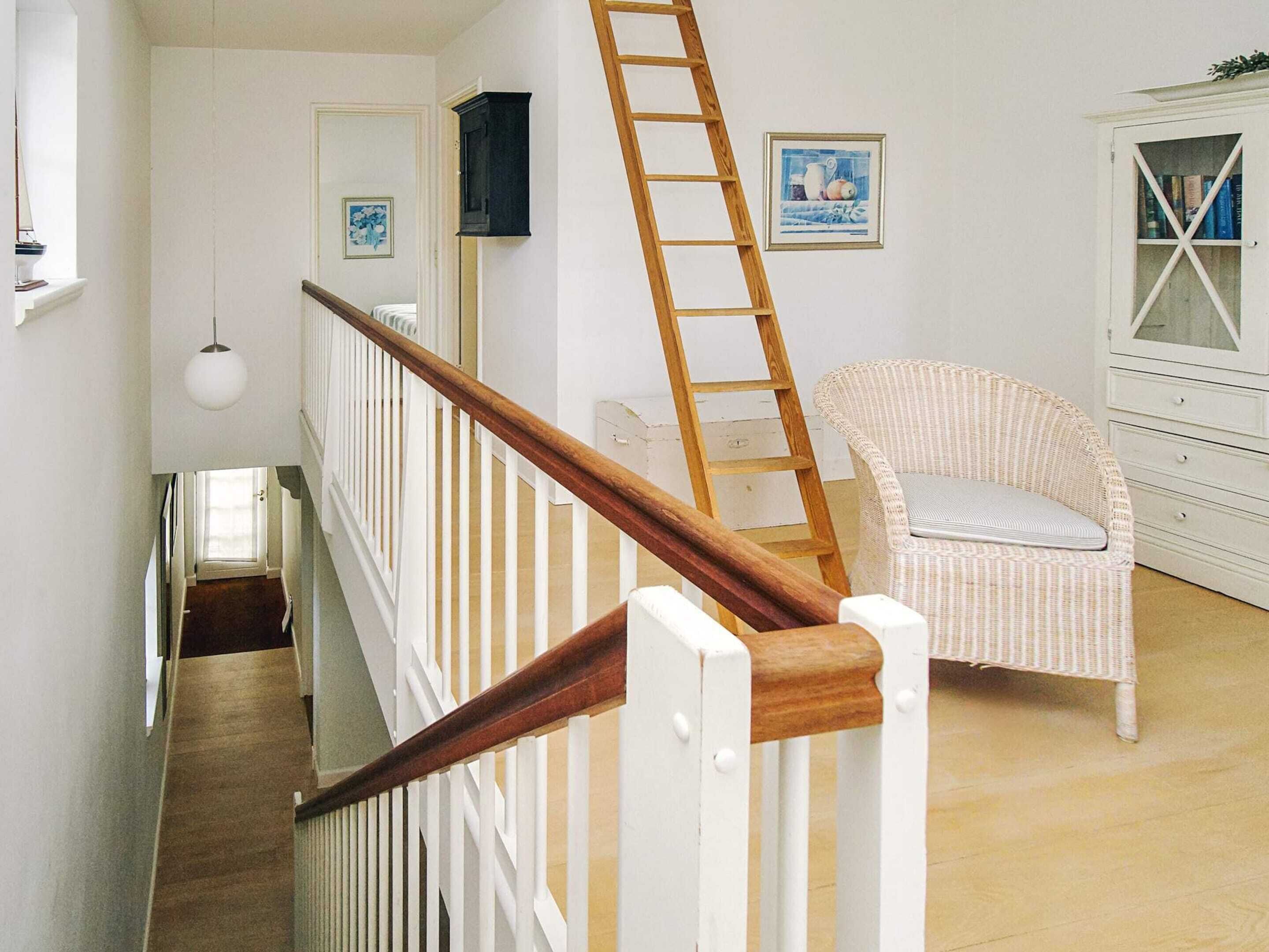 Una escalera con barandilla de madera y balaustres blancos, una silla de mimbre con cojín, un armario blanco con puertas de cristal y un cuadro en la pared.