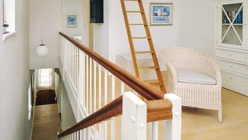 Una escalera con barandilla de madera y balaustres blancos, una silla de mimbre con cojín, un armario blanco con puertas de cristal y un cuadro en la pared.