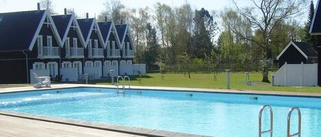 A swimming pool with a wooden deck, a building with a dark roof and white trim, and trees in the background.